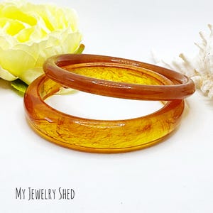 May include: Two amber-colored bangle bracelets with a marbled, swirled pattern. The bracelets are stacked on top of each other.