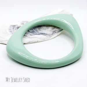 May include: A light green, glossy,  smooth,  and  curved bangle bracelet.  The bracelet is made of plastic and has a unique, modern design.