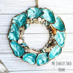 May include: A circular wreath made of turquoise-colored oyster shells with gold edges, interspersed with various seashells and pearls. The wreath is suspended by a natural twine loop. The text "MY JEWELRY SHED HOME" is visible.