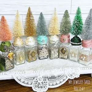 May include: A collection of decorative glass jars, each adorned with a miniature bottle brush tree in various colors. The jars are embellished with ribbons, jewels, and vintage-style ornaments, creating a festive holiday display. The jars are arranged on a white tray with a lace trim.