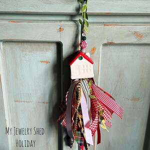 May include: A festive holiday ornament featuring a white ceramic house with a red roof, green accents, and a tassel of ribbons in red, white, and green patterns. The ornament is suspended from a green vine.