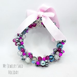 May include: A beaded holiday wreath with a pink satin ribbon bow. The wreath is made of various sized beads in shades of pink, purple, silver, and iridescent blue. The text "My Jewelry Shed Holiday" is at the bottom.