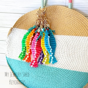 May include: A collection of colorful beaded fish keychains in various colors, including green, orange, pink, red, yellow, purple, and blue. The keychains are attached to a gold ring. The background is a round straw bag with white, blue, and tan stripes. Text on the bag reads "My Jewelry Shed Keychains."