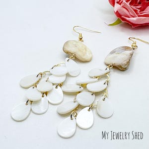 May include: White shell earrings with gold accents. The earrings feature a large shell charm and a cascading design of smaller shell charms.