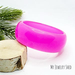 May include: A vibrant, hot pink bangle bracelet. The chunky, rounded bracelet is made of a glossy material. The bracelet is resting on a piece of wood and a sprig of evergreen. The text "MY JEWELRY SHED" is in the lower right corner.