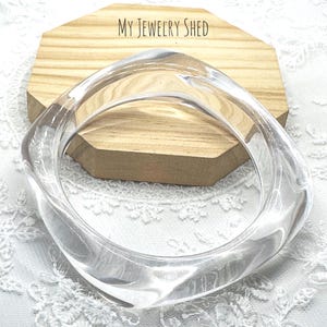 May include: A clear, transparent bangle bracelet with a smooth, rounded square shape. The bracelet is made of a solid, glass-like material that reflects light, creating subtle highlights and shadows along its curved, polished surface.