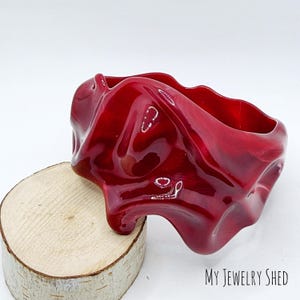 May include: A deep red, glossy bangle bracelet with an irregular, undulating, and organic shape. The surface of the bracelet is smooth and reflective, catching light across its curved, flowing form. The text "MY JEWELRY SHED" is visible.