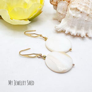 May include: A pair of gold-colored earrings with white, oval-shaped pendants. The earrings have a simple hook design and small gold beads. The background includes a yellow flower, a seashell, and the text "MY JEWELRY SHED".