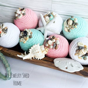 May include: A collection of six decorative rope balls, each wrapped in white or light blue and pink rope. The balls are decorated with seashells and pearls. The balls are arranged in a wooden bowl with a white seashell.
