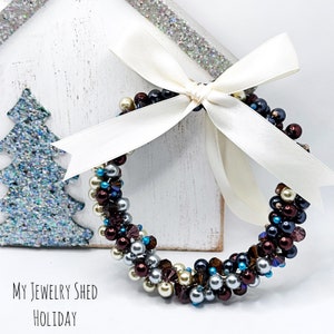May include: A beaded wreath with a cream-colored ribbon bow. The wreath is made of various colored beads, including silver, burgundy, blue, and pearl. The text "My Jewelry Shed Holiday" is at the bottom.