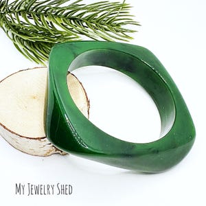 May include: A square-shaped, dark green bangle bracelet. The bracelet has a glossy finish and a geometric design. The image also includes a sprig of greenery and a piece of wood. The text "MY JEWELRY SHED" is visible at the bottom of the image.