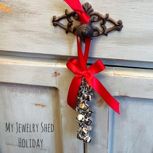 May include: A cluster of silver bells tied with a bright red ribbon, hanging from a decorative, dark metal handle. The ribbon is tied in a bow. The text "MY JEWELRY SHED HOLIDAY" is visible in the lower left corner.