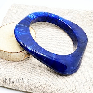 May include: A square-shaped blue bangle bracelet with a marbled effect. The bracelet is made of a glossy, translucent material. The words "MY JEWELRY SHED" are printed in black at the bottom of the image.