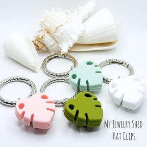 May include: A collection of hat clips in the shape of Monstera leaves, in pink, green, white, and light blue, attached to silver key rings. The text "MY JEWELRY SHED HAT CLIPS" is visible in the image. Decorative seashells are in the background.