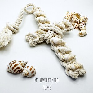 May include: A white rope cross with seashells woven into the rope. The cross has a tassel at the top. There are also seashells scattered around the cross.