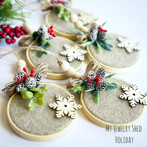 May include: Circular Christmas ornaments with a burlap-like center, wooden frame, and decorative elements. Each ornament features a small bundle of pinecones, red berries, and greenery, tied with twine. Wooden snowflake accents and the text "My Jewelry Shed Holiday" are also visible.