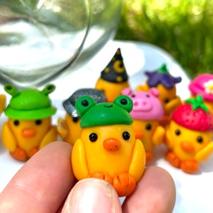 May include: A collection of yellow chick figurines wearing various hats, including a green frog hat, a pink pig hat, a strawberry hat, and a witch hat.