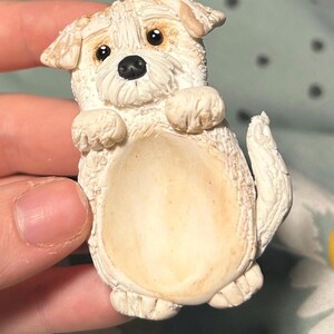 Puede incluir: Una figurita de cer&aacute;mica blanca de un perro peque&ntilde;o con ojos marrones y nariz negra. El perro est&aacute; de pie sobre sus patas traseras con las patas delanteras levantadas.
