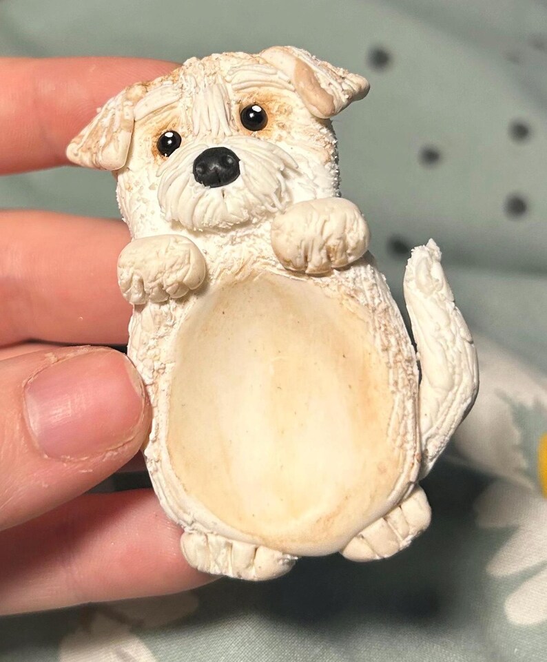 Puede incluir: Una figurita de cer&aacute;mica blanca de un perro peque&ntilde;o con ojos marrones y nariz negra. El perro est&aacute; de pie sobre sus patas traseras con las patas delanteras levantadas.