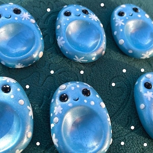 May include: Six blue, oval-shaped ring holders with smiling faces and white floral and dot accents. The ring holders have a recessed centre for holding rings. The background is a teal green colour.