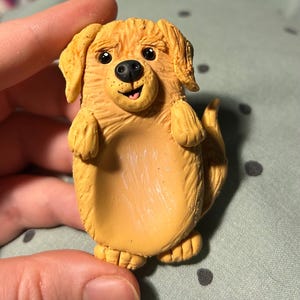 Puede incluir: Una figurita de perro golden retriever hecha a mano de arcilla polim&eacute;rica. El perro est&aacute; de pie sobre sus patas traseras con las patas delanteras levantadas. Tiene una expresi&oacute;n feliz con una amplia sonrisa y la lengua fuera.