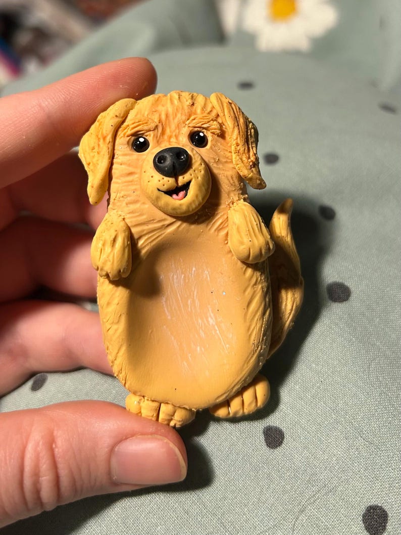 Puede incluir: Una figurita de perro golden retriever hecha a mano de arcilla polim&eacute;rica. El perro est&aacute; de pie sobre sus patas traseras con las patas delanteras levantadas. Tiene una expresi&oacute;n feliz con una amplia sonrisa y la lengua fuera.