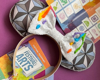 May include: White sparkly Minnie Mouse ears with a rainbow paint splatter design. The ears are shaped like the Epcot Spaceship Earth and feature a silver trim. The headband is brown. The image also includes a pamphlet for the Epcot International Festival of the Arts.
