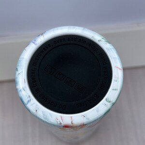 May include: White ceramic tumbler with a floral design. The bottom of the tumbler has a black lid with the text "BPA free Do not use in freezer, microwave, oven and stovetop. Hand wash only".