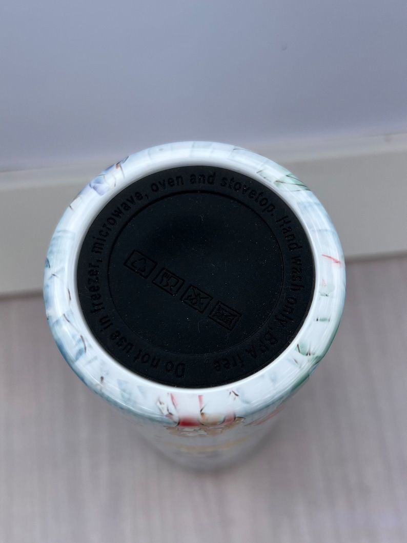 May include: White ceramic tumbler with a floral design. The bottom of the tumbler has a black lid with the text "BPA free Do not use in freezer, microwave, oven and stovetop. Hand wash only".