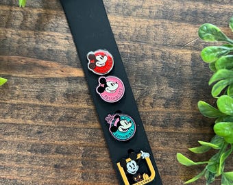 Puede incluir: Una banda de reloj de silicona negra con cuatro pines de portador de pase de Mickey Mouse de colores adjuntos. Los pines son redondos y presentan diferentes poses de Mickey Mouse. El texto "Passholder" está impreso en cada pin.