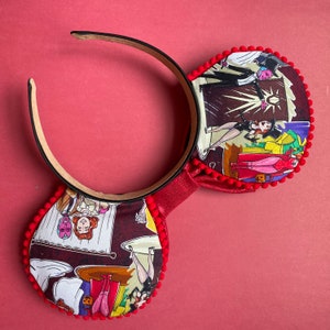 May include: A pair of mouse ears with a red headband and ear details. The ears are decorated with a colorful comic strip pattern and trimmed with red pom-poms. The bow is red and textured. The background is a solid red.