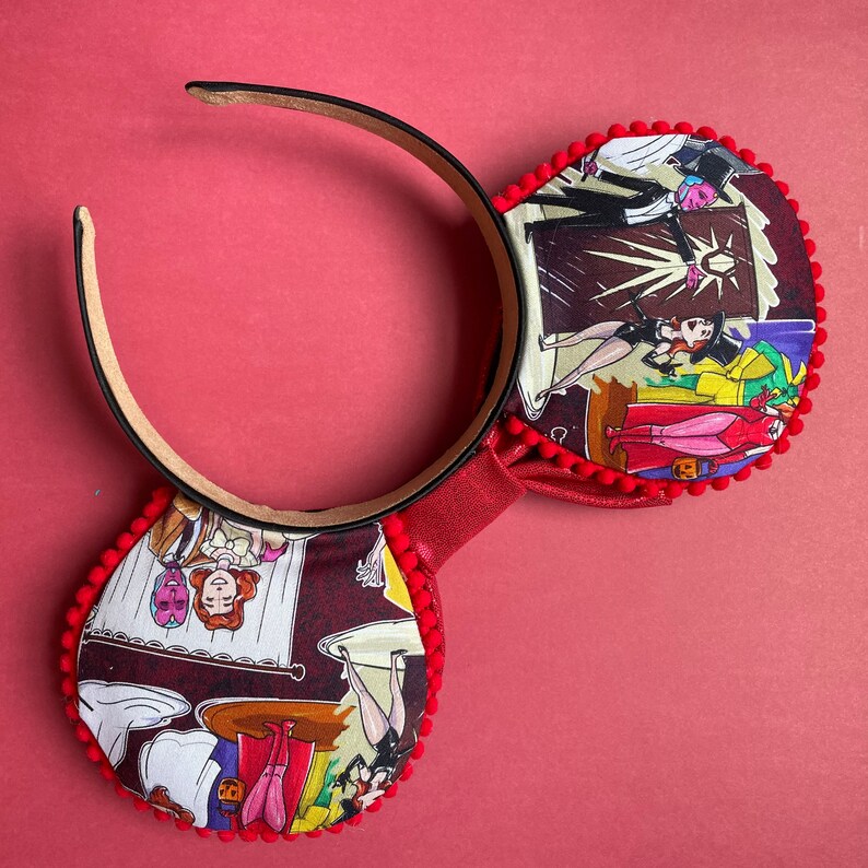 May include: A pair of mouse ears with a red headband and ear details. The ears are decorated with a colorful comic strip pattern and trimmed with red pom-poms. The bow is red and textured. The background is a solid red.