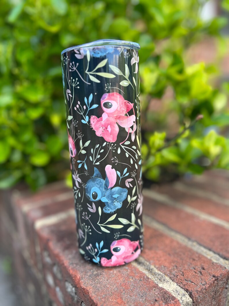 May include: A black stainless steel tumbler with a floral pattern featuring pink and blue cartoon characters. The tumbler has a clear lid.