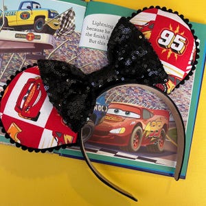 May include: A black sequined bow headband with Cars-themed ear details. The ears feature Lightning McQueen and the number 95 on a red and white checkered background. The headband is resting on an open book with illustrations of the Cars movie.