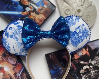 Puede incluir: Orejas de ratón de tela con estampado azul y blanco con un lazo de lentejuelas azules. Las orejas están rodeadas de carteles de películas de Star Wars.