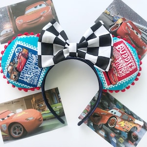 May include: A pair of blue and red mouse ears with a black and white checkered bow. The ears feature images of Lightning McQueen and Mater from the Disney Pixar Cars movie. The text on the ears reads "Lightning McQueen, Tow Mater, Fuelin' Up for the Race!" and "Buddies to the Finish".