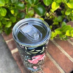 May include: A black and white tumbler with a floral design featuring pink, blue, and green flowers. The tumbler has a clear lid with a slider.