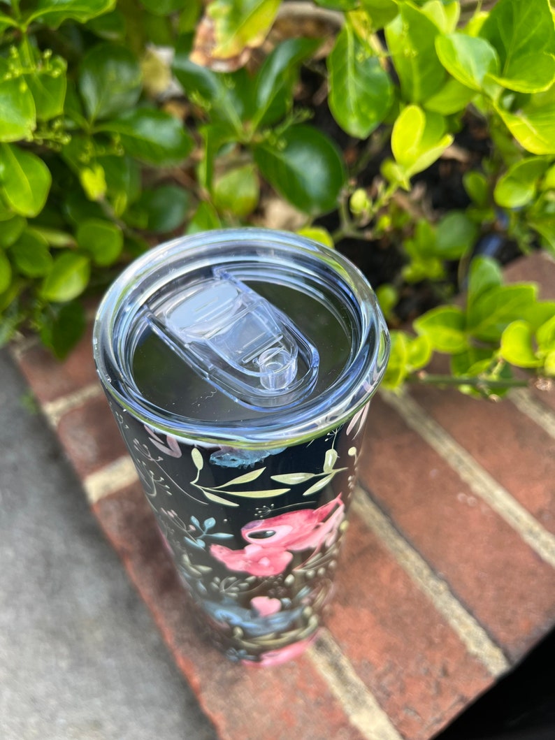 May include: A black and white tumbler with a floral design featuring pink, blue, and green flowers. The tumbler has a clear lid with a slider.