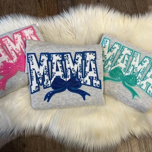 May include: Three gray sweatshirts with the word "MAMA" in white floral lettering and a bow. One sweatshirt has pink accents, one has blue, and one has teal. The sweatshirts are displayed on a white fluffy surface.