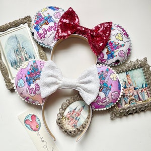 May include: A pair of white mouse ears with a pink and purple fabric design featuring a castle and the word "home". The ears are decorated with a white bow and a pink and red sequined bow.
