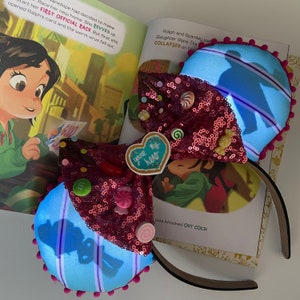 Peut inclure: Oreilles de Minnie Mouse roses et bleues avec un nœud rose pailleté et un charme en forme de cœur qui dit "you're my hero". Les oreilles sont décorées de bonbons et d'autres friandises.