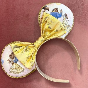 Könnte beinhalten: Gelbe und weiße Stoff-Mausohren mit goldener Einfassung. Die Ohren zeigen einen Aufdruck von Belle und dem Biest aus Disneys Die Schöne und das Biest.
