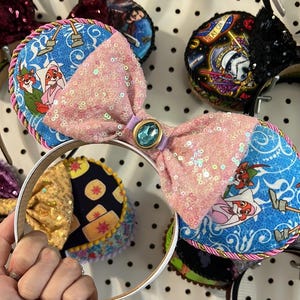 May include: A selection of decorative headbands with diverse designs. One headband displays a pink sequined bow with a blue gem, and blue ear details with cartoon characters. Other headbands feature different patterns and colours.
