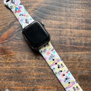 May include: Black smartwatch with a white band featuring a colorful cartoon design. The band has images of cartoon characters, castles, and fireworks. The watch is on a wooden surface.