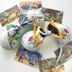 May include: A pair of white and blue Toy Story themed Minnie Mouse ears with a cow print bow. The ears feature images of Woody and Buzz Lightyear. The bow is made of a yellow and white checkered fabric with a black and white cow print.
