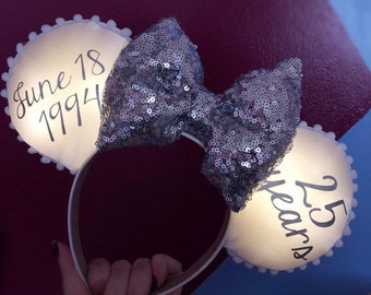 May include: White light-up Minnie Mouse ears with a silver sequined bow. The left ear says "June 18 1994" and the right ear says "25 Years".