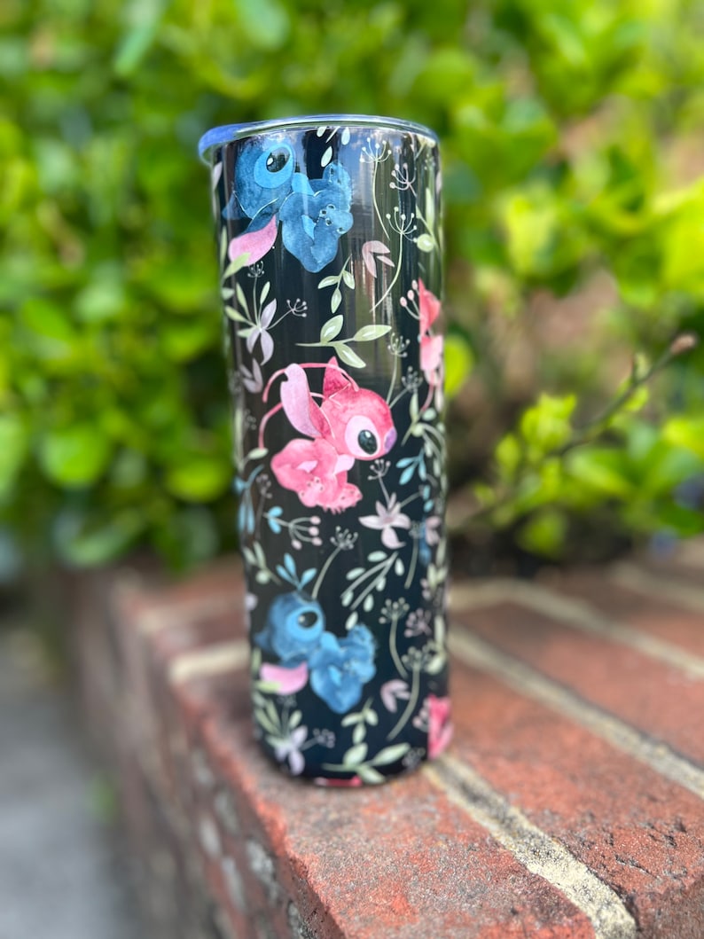 May include: A black stainless steel tumbler with a floral pattern and cartoon characters. The characters are blue, pink, and white. The tumbler has a lid.