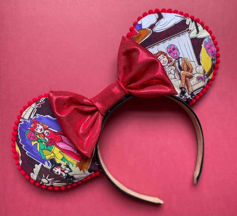 May include: Red Minnie Mouse ear headband with a large, shiny red bow. The ears feature a comic strip design with characters in red, pink, and purple. The ears are trimmed with red pom-poms, and the headband is black.