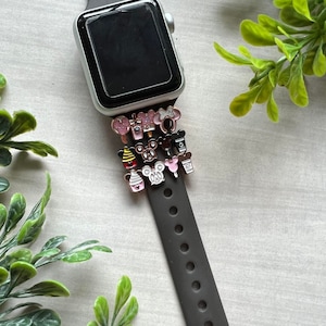 May include: A black Apple Watch with a black band. The band is decorated with small, colorful enamel charms featuring Disney characters and food items.