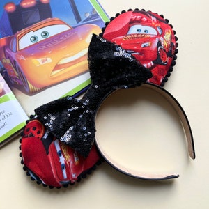 May include: Red and black mouse ears with a black sequin bow. The ears feature a printed image of Lightning McQueen from the Disney Pixar Cars movie.